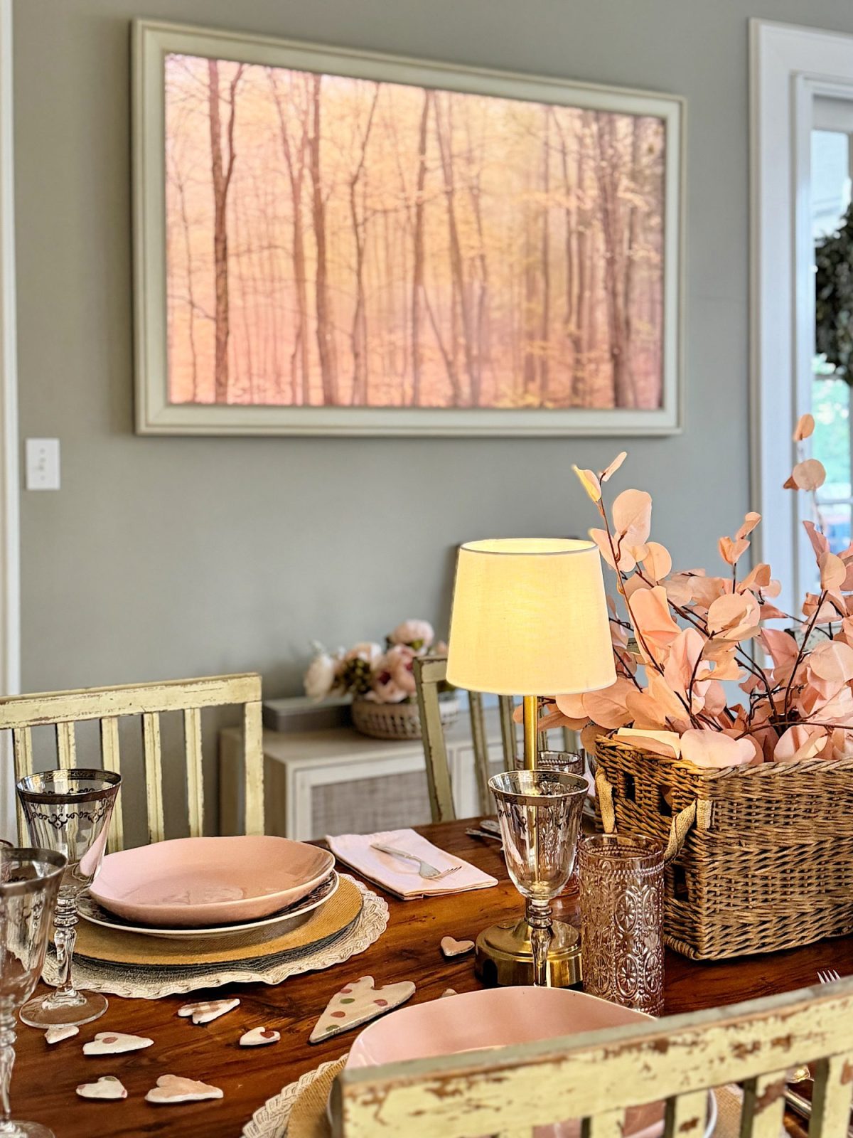 Dining area with a wooden table set with pink plates, glasses, and a lamp. A woven basket with pink leaves decorates the center. A framed tree landscape image hangs on the wall.