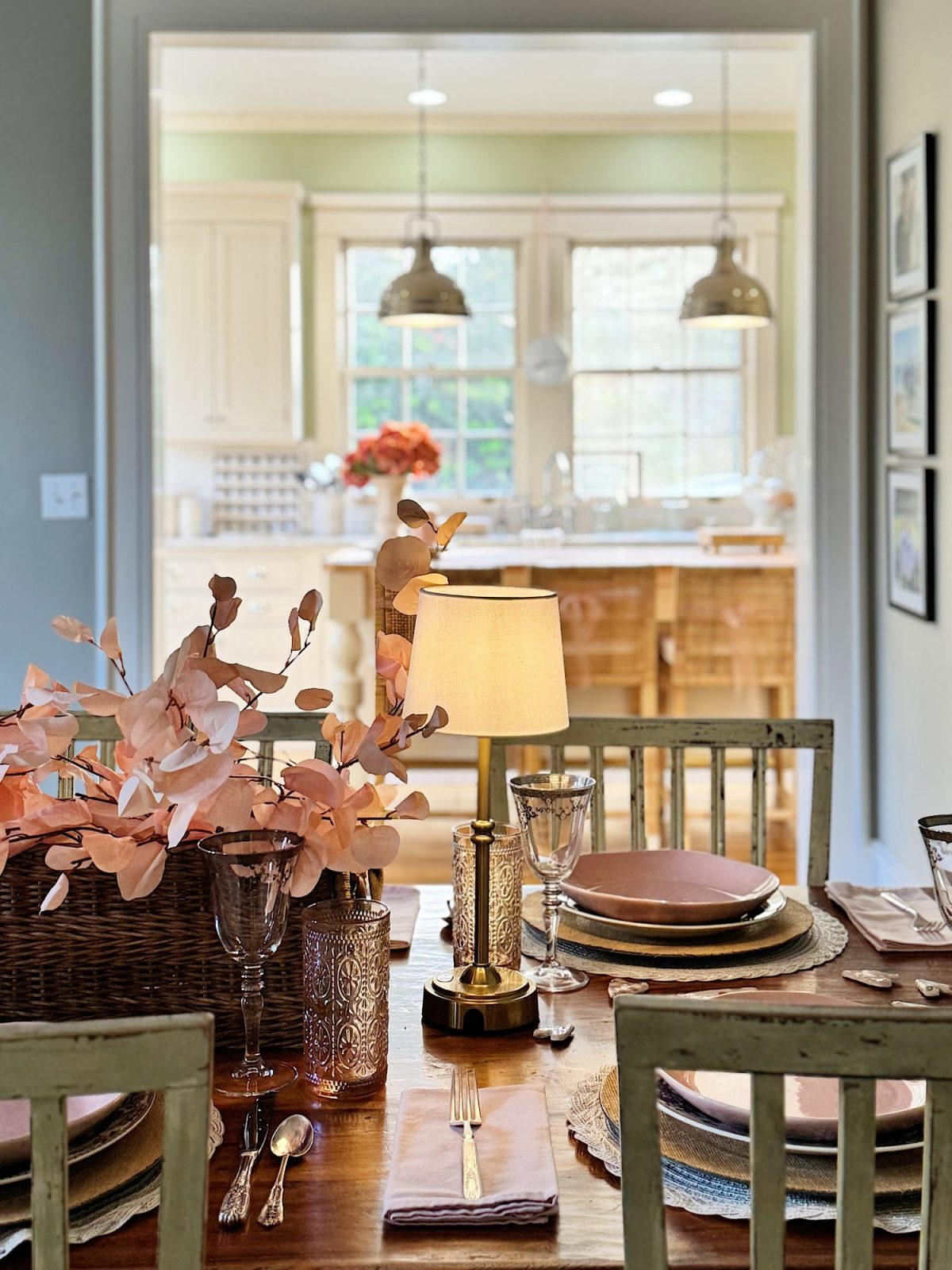 A dining table set with a lamp, pink napkins, and glassware. In the background, a kitchen with hanging lights, wooden island, and a basket with pink flowers can be seen.