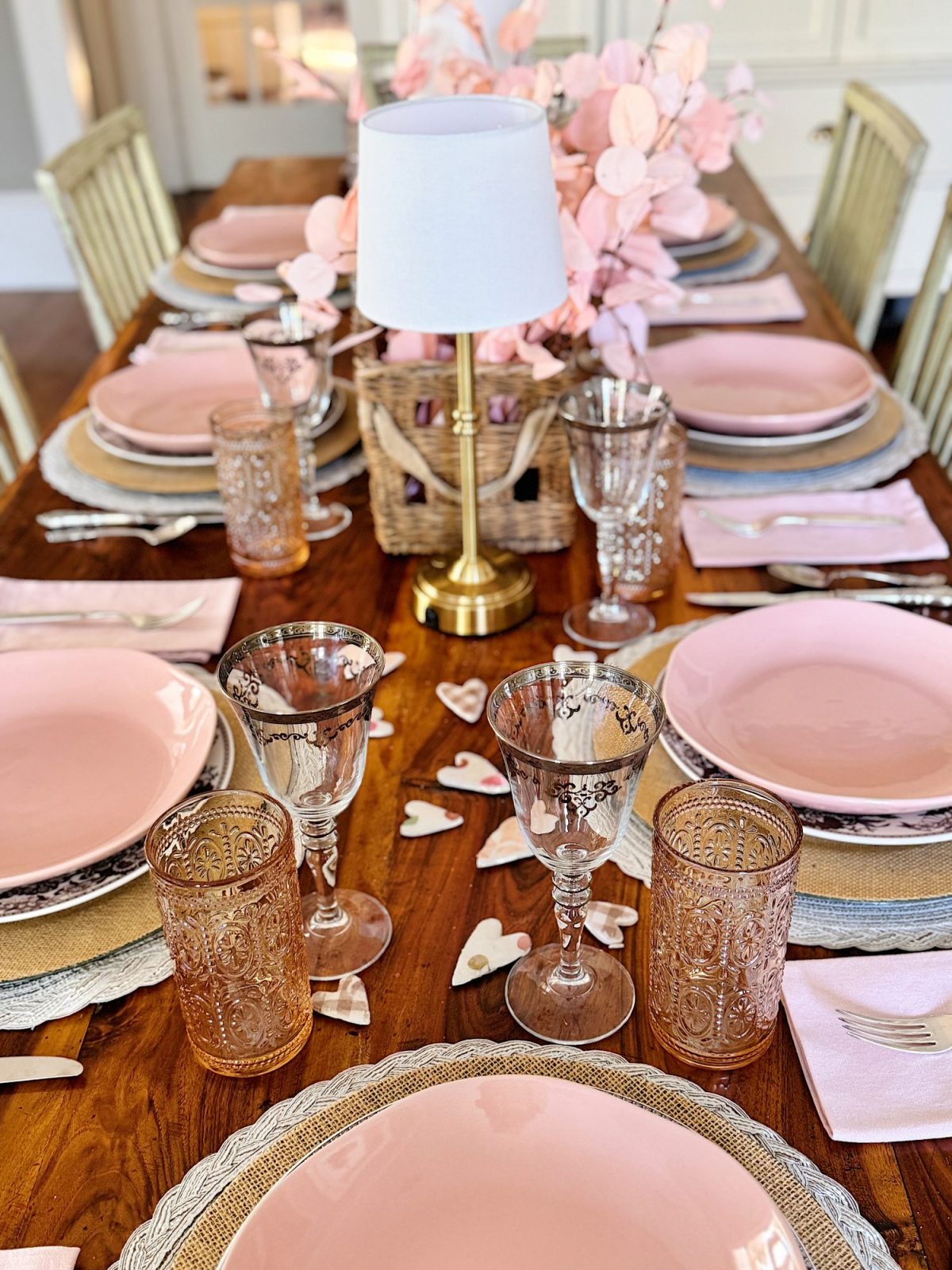 A dining table set with pink plates, ornate glasses, and napkins. A lamp with a pink floral centerpiece is in the middle.