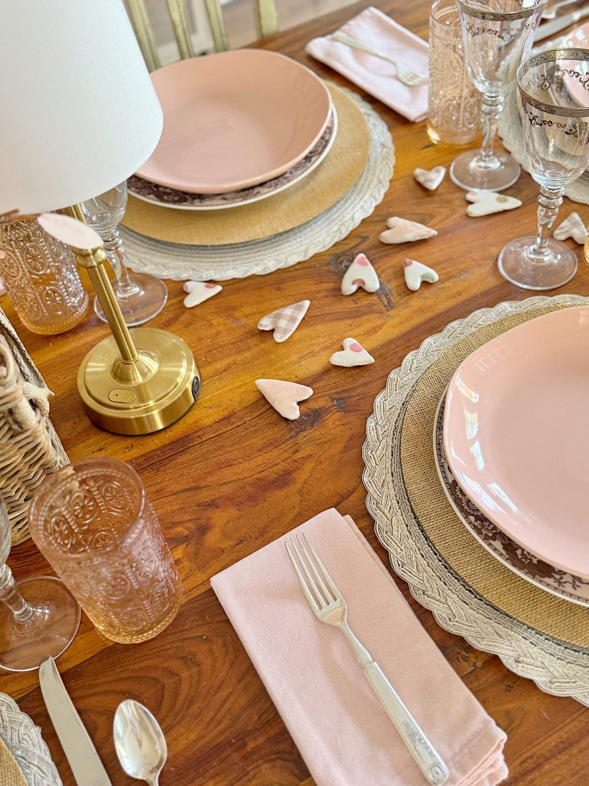 A dining table set for two with pink plates, glasses, and napkins on woven placemats. Decorative heart-shaped items are scattered on the wooden surface. A small lamp sits at the table's corner.