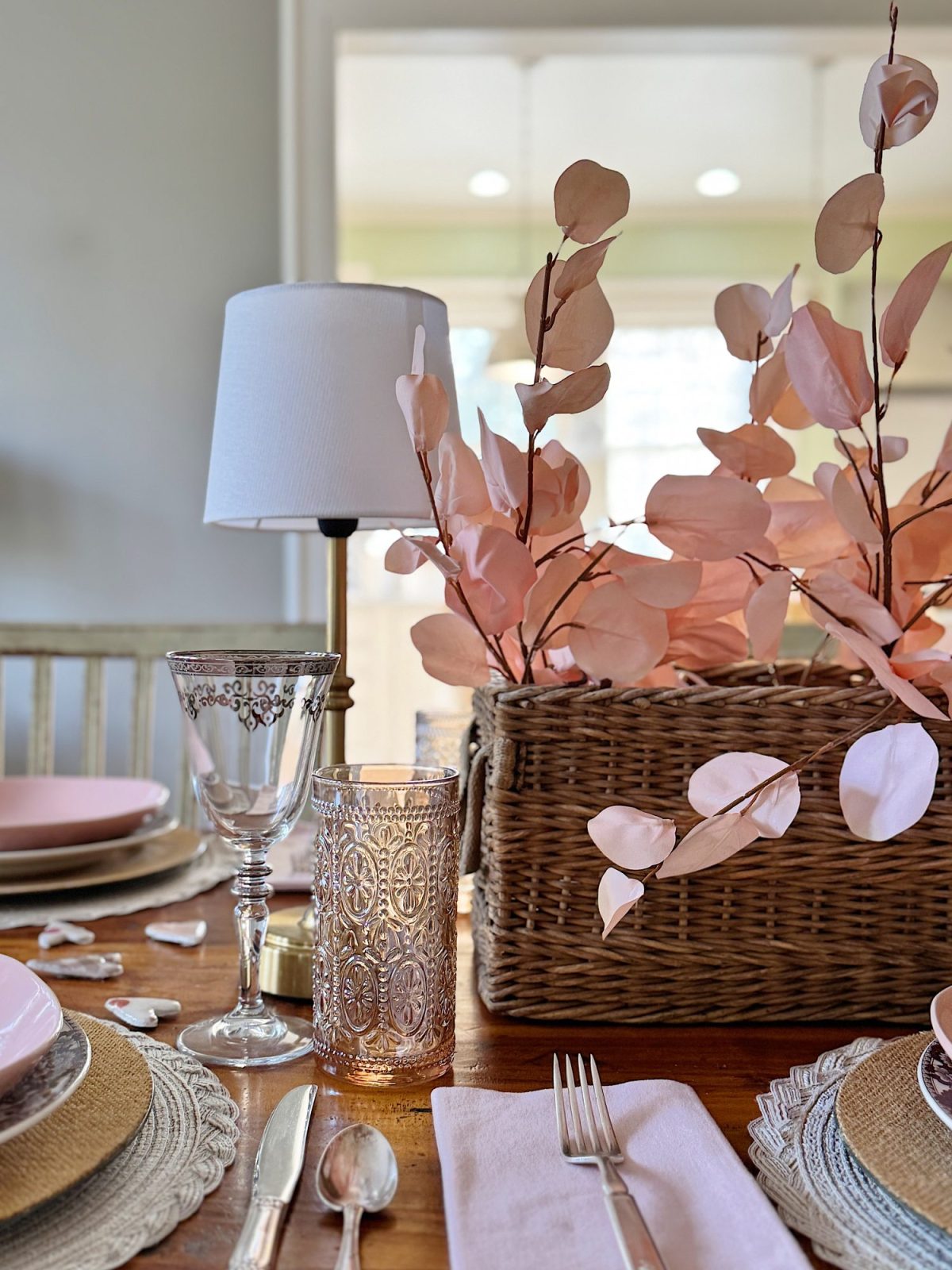 Table setting with a woven basket holding pink decorative leaves. Includes a wine glass, candle holder, and cutlery on a wooden table with pink and beige tableware and a small lamp.