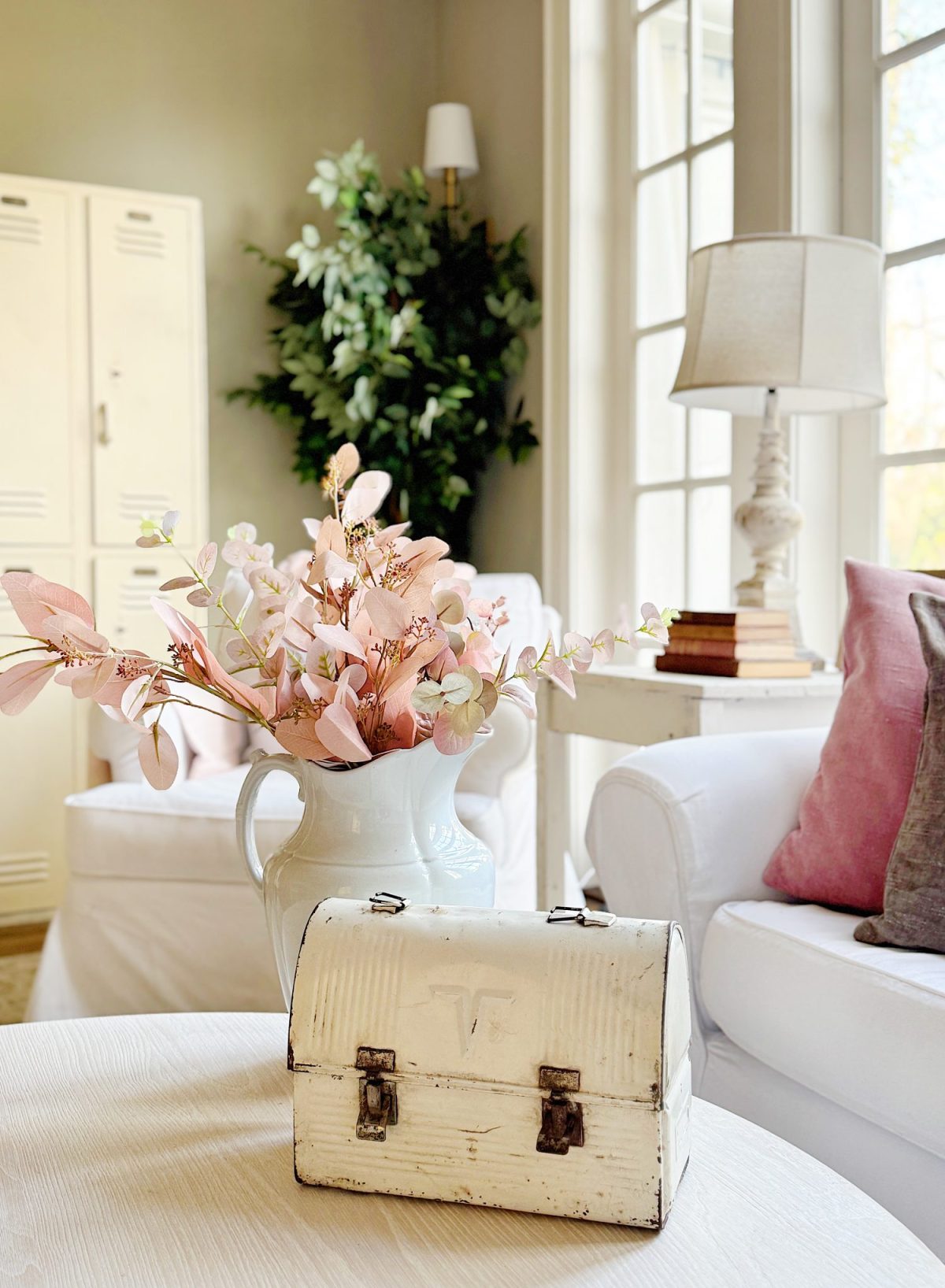 A white metal lunchbox on a round table in a living room with a white armchair, a large plant, and a pitcher with pink flowers. A lamp and stacked books are beside the armchair.
