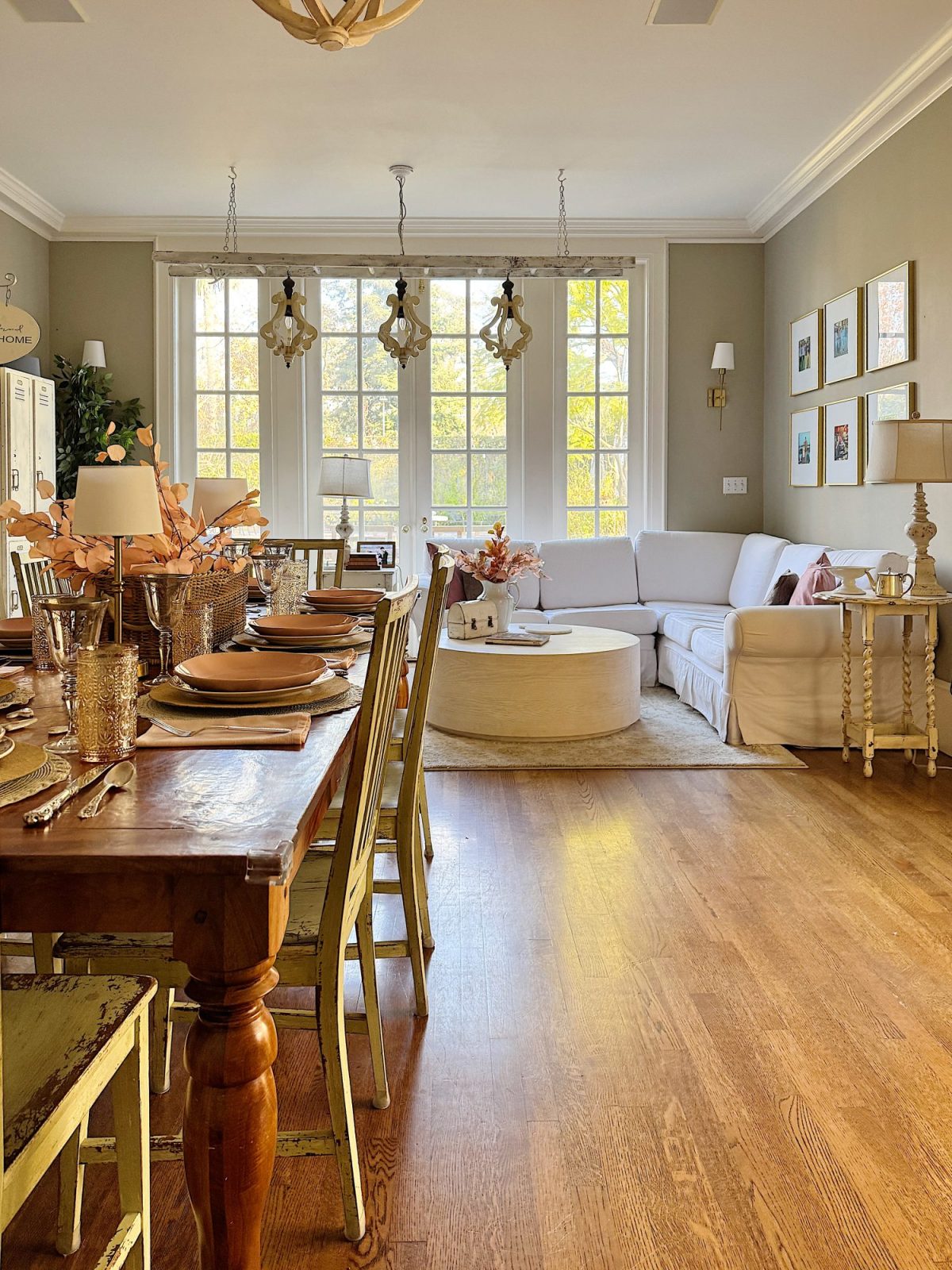 A cozy living and dining space with a wooden table, chairs, and place settings on the left. A white couch, round coffee table, and large windows with hanging lights on the right.