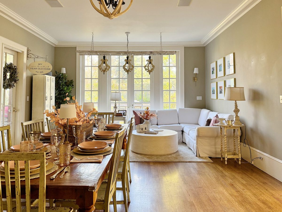 A cozy dining and living room features a wooden dining table set with dishes, a white sectional sofa, a round coffee table, large windows, light walls, and decorative lighting.