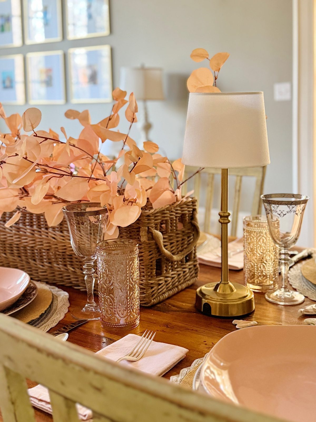 A dining table set with pink plates, decorative glasses, a small lamp, and a wicker basket filled with peach-colored leaves.