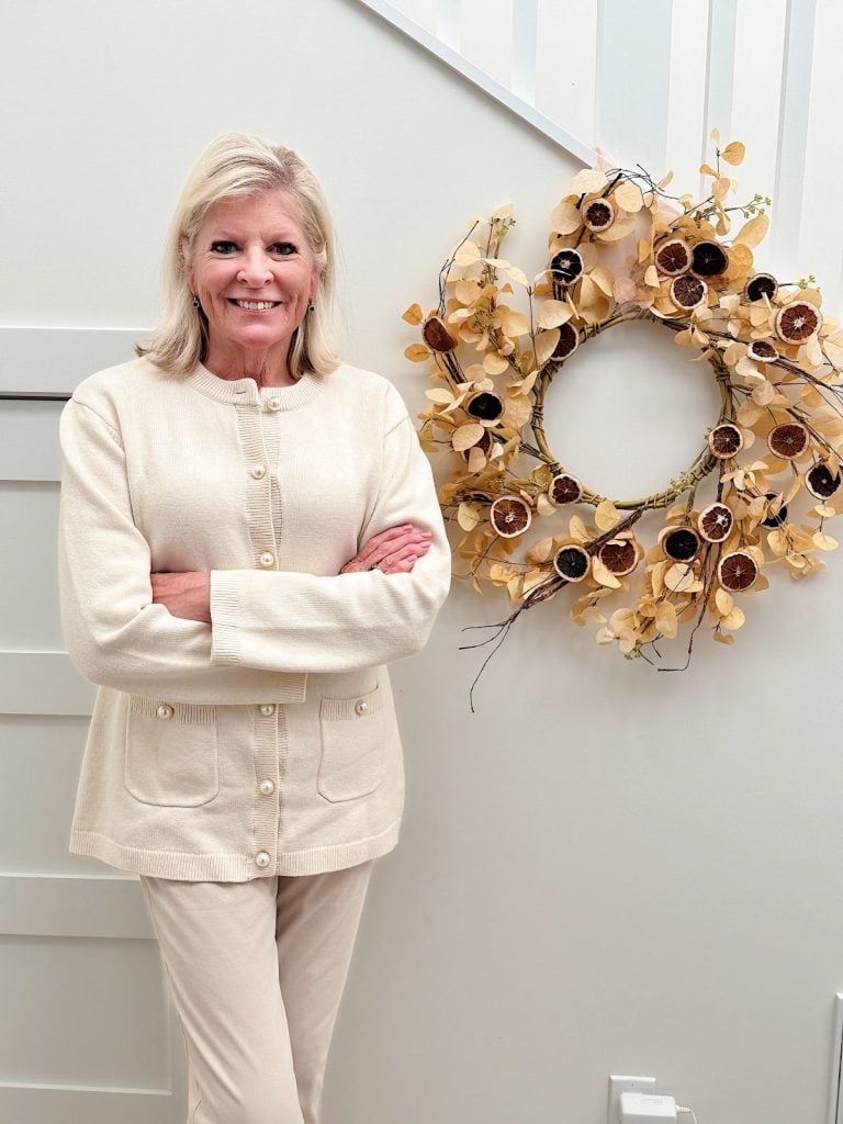 A person stands smiling with arms crossed next to a decorative autumn wreath on a light-colored wall, embracing the festive spirit of both fall and Happy New Year.