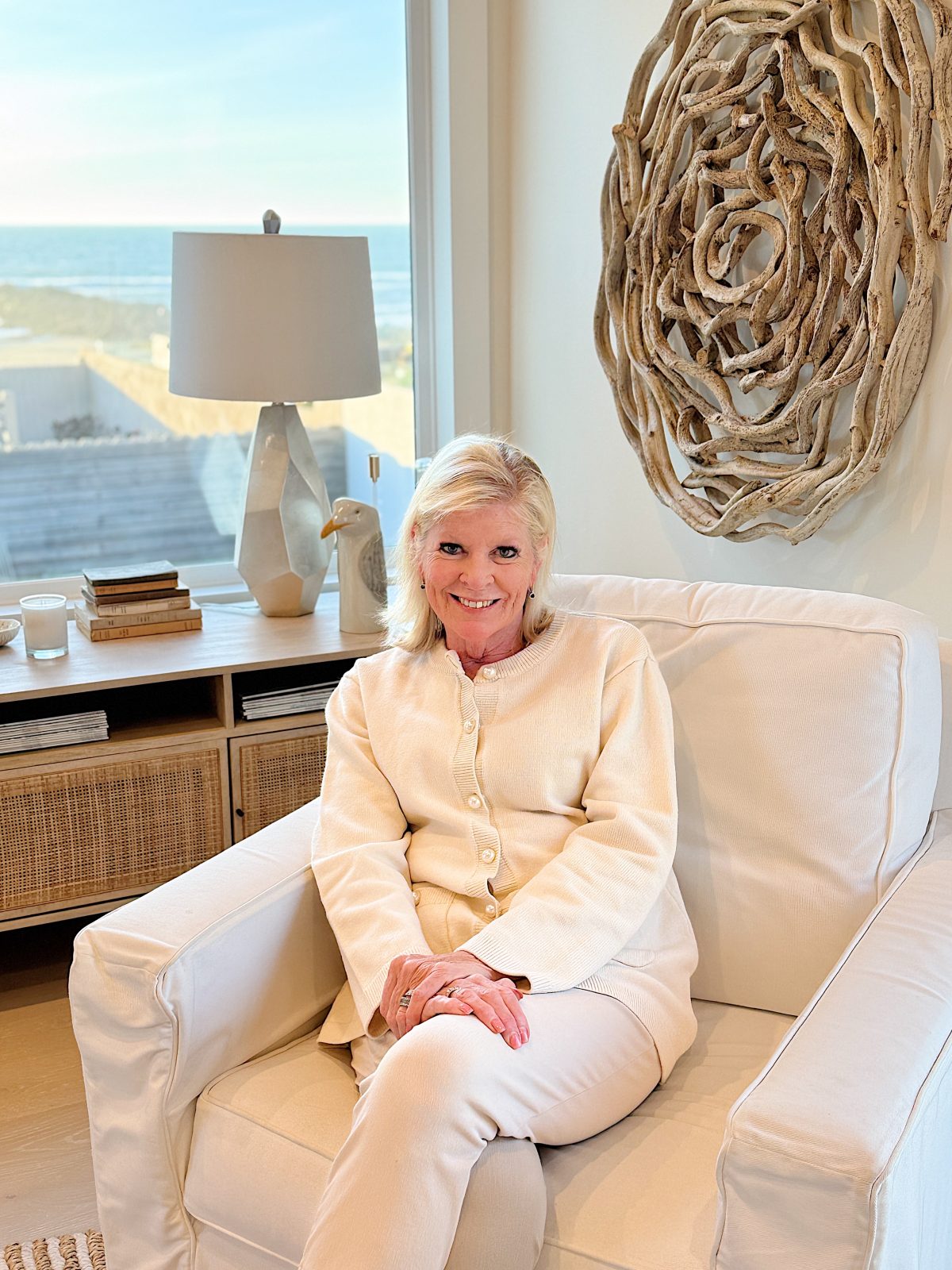 A person sitting on a white armchair in a bright room with a large window, a decorative wood wall piece, and a lamp in the background, enjoying the peaceful ambiance and reflecting on new beginnings as they prepare to celebrate Happy New Year.