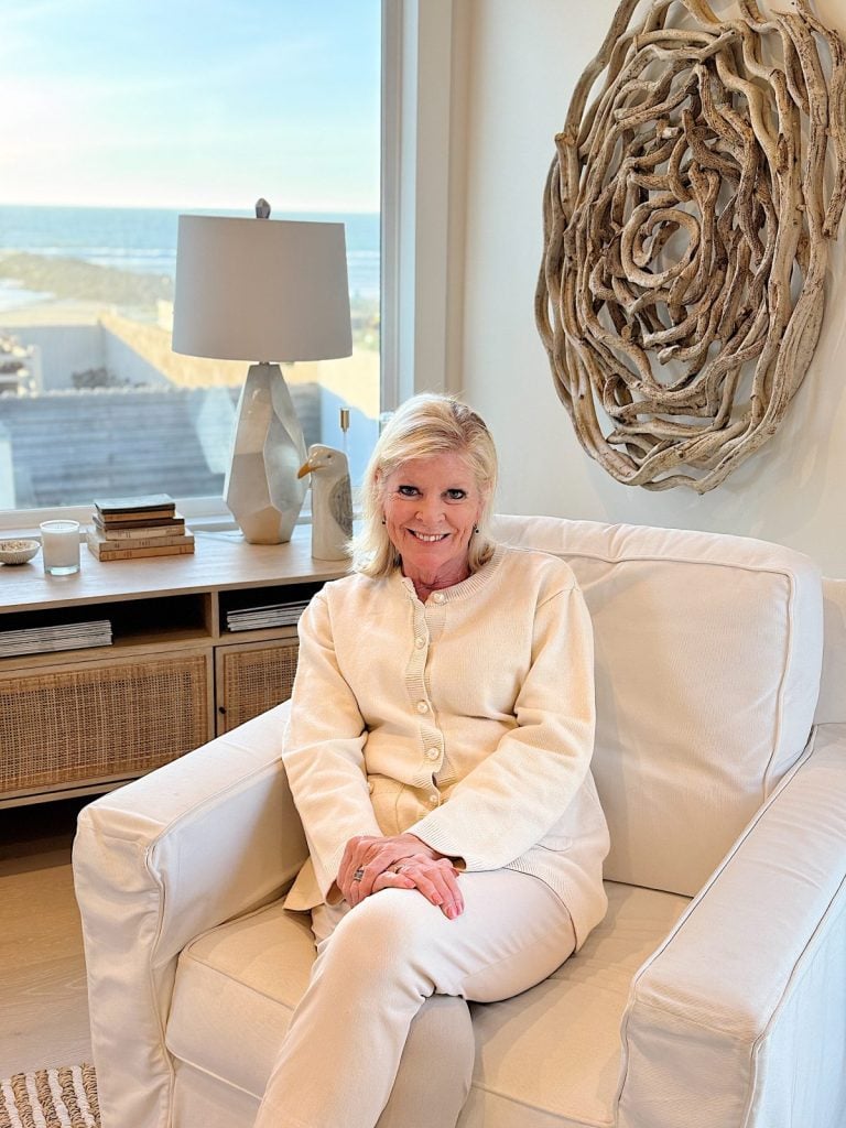 A woman in a cream outfit sits in an armchair near a window with a beach view, embracing the serene ambiance. Behind her, driftwood wall art and a lamp complete the cozy setting, as she quietly reflects on the Happy New Year ahead.