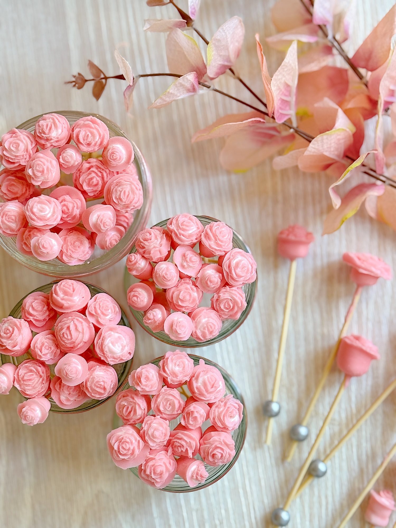 Glass jars filled with small pink rose-shaped candies on a wooden surface, with pink faux flowers and skewers nearby.