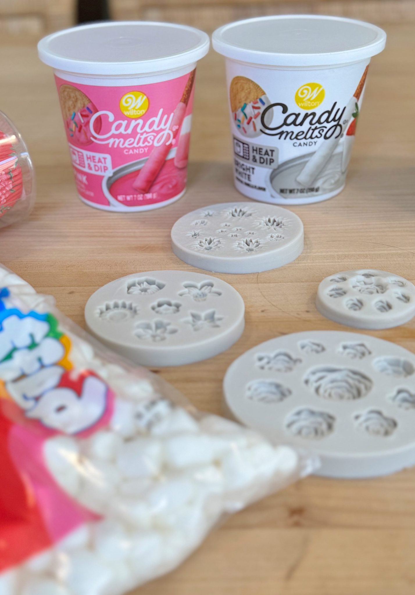Plastic tubs of candy melts and silicone molds on a wooden surface, with a bag of marshmallows in the foreground.