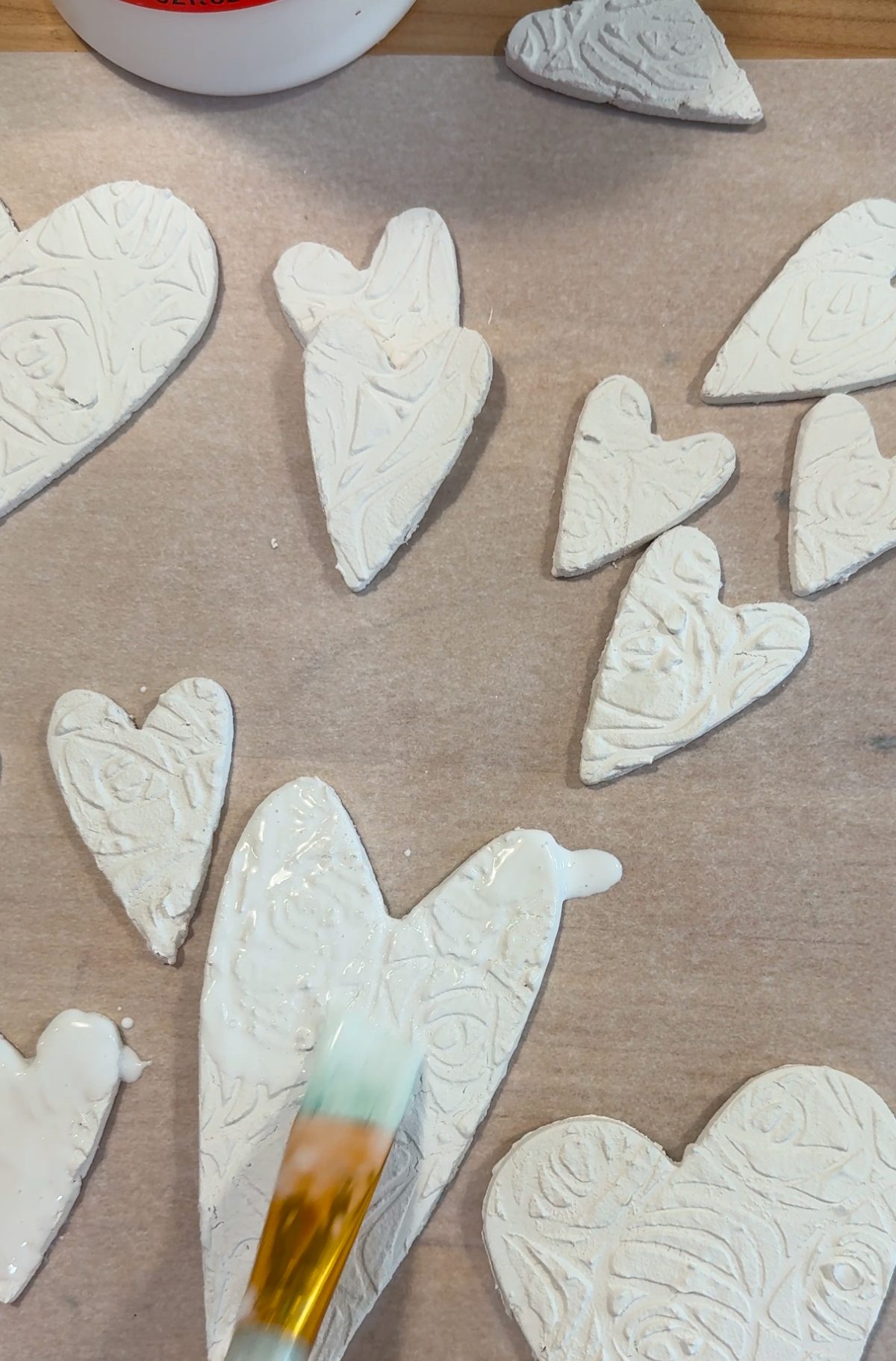 A brush applies glue to heart-shaped clay pieces with patterns.
