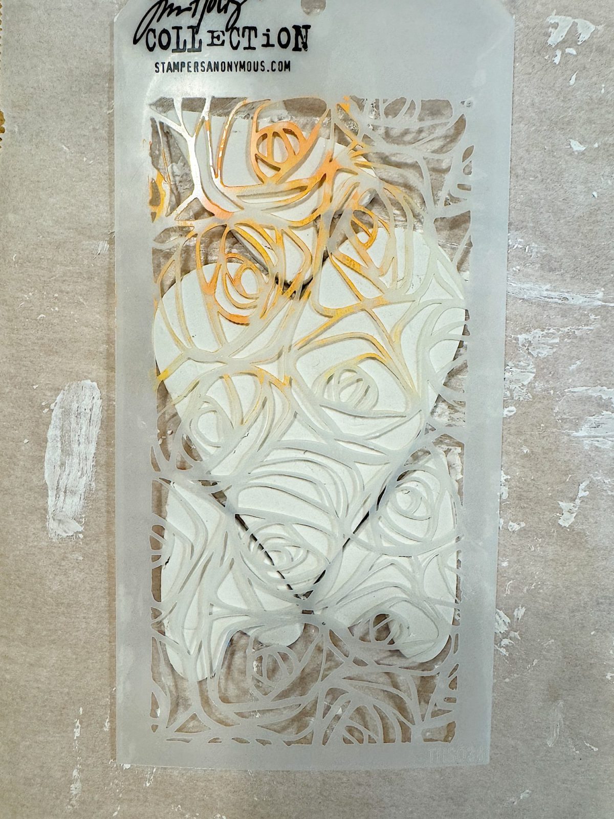 A plastic stencil with a rose pattern design is placed on a textured surface. The stencil has some areas marked with orange ink.