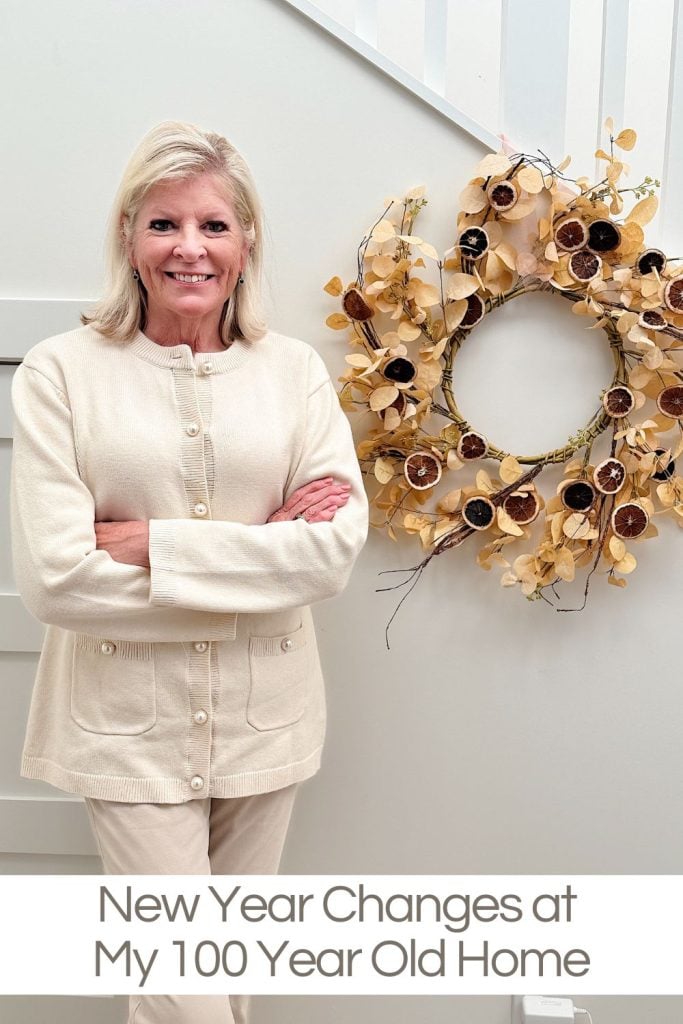 Smiling Leslie Saeta in a light sweater stands with arms crossed next to a decorative wreath on a wall. Text reads, "New Year Changes at My 100 Year Old Home.