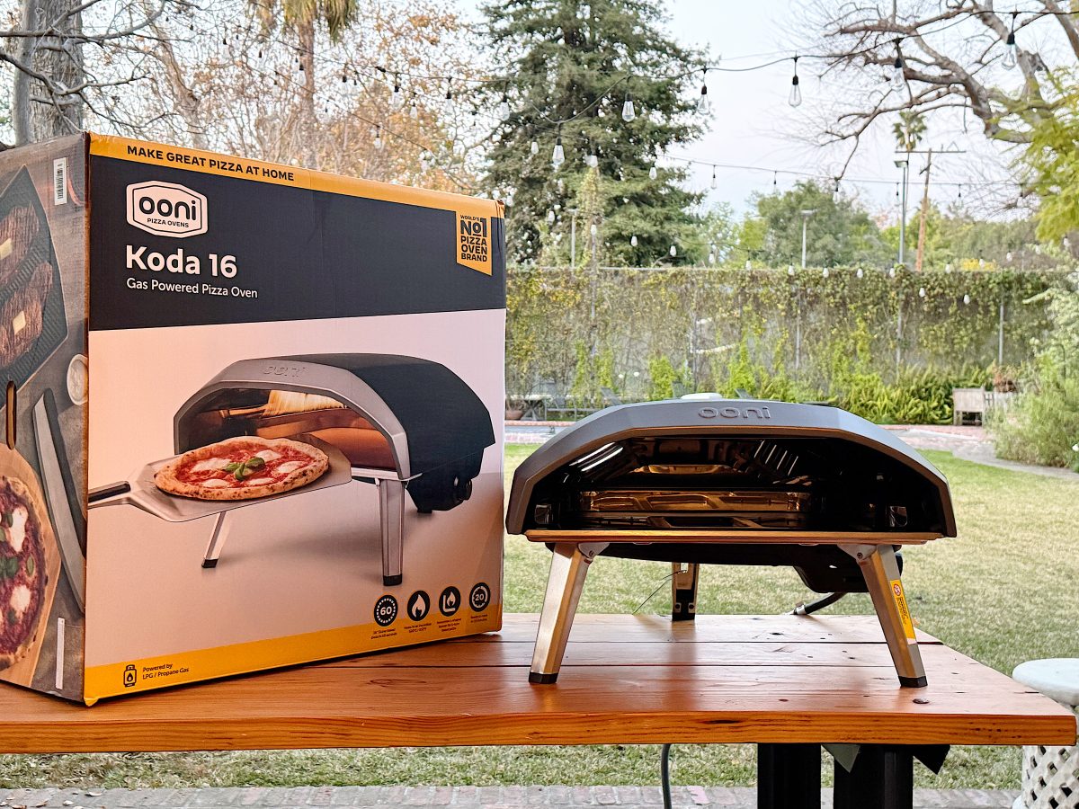 Portable pizza oven on a wooden table outside, next to its box labeled "Ooni Koda 16 Gas Powered Pizza Oven." Trees and greenery in the background.