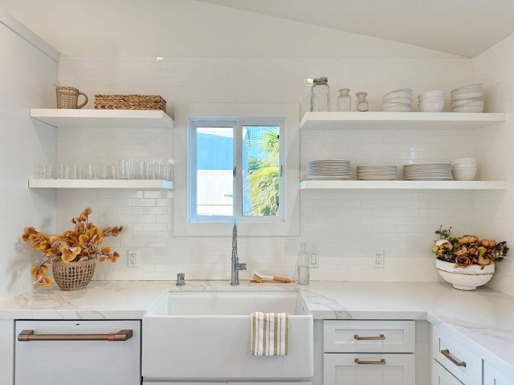 A bright kitchen features a farmhouse sink, white cabinets, open shelves with plates and glasses, and decorative baskets with dried flowers. A window above the sink lets in natural light.