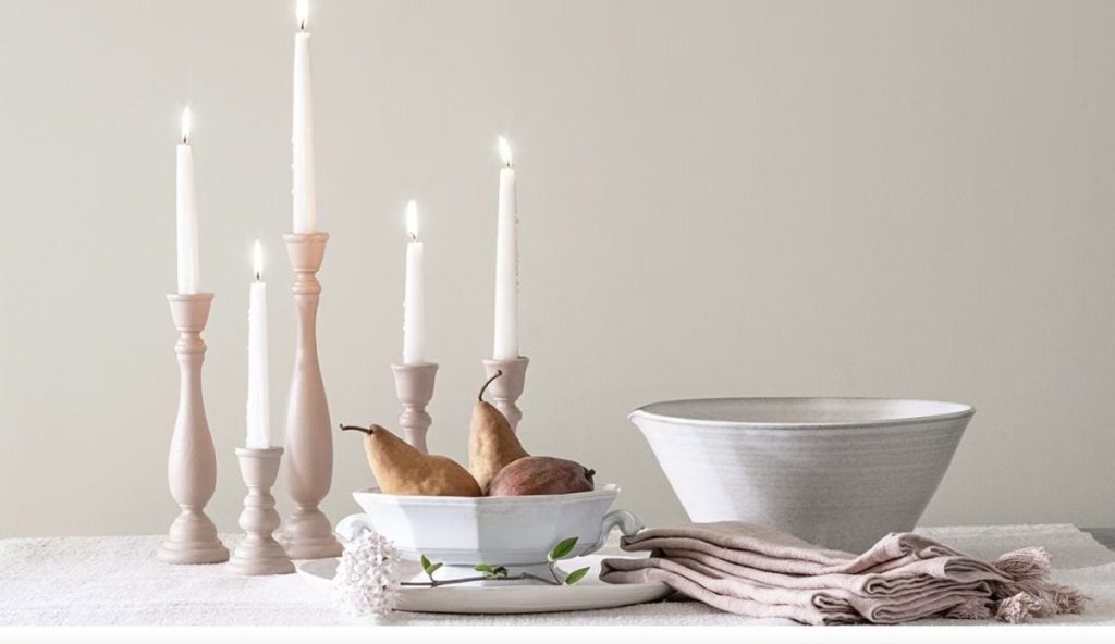 A still life scene with lit candles, a bowl with pears and an onion, a large ceramic bowl, folded cloth, and a small flower on a light-colored surface embodies the serene essence of a winter color palette.