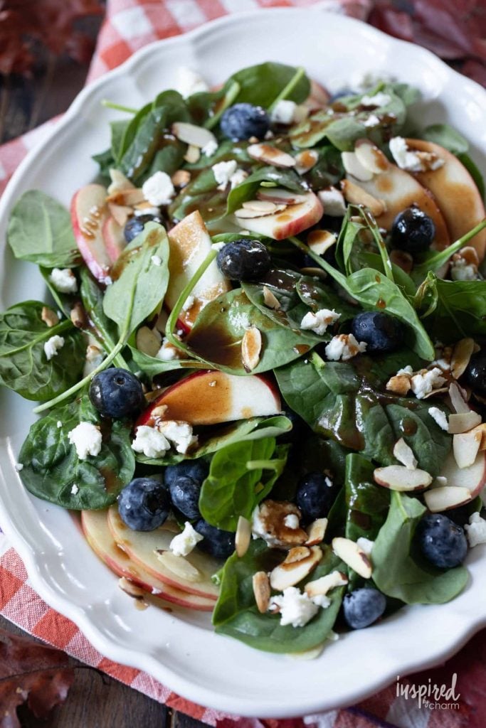 A plate of spinach salad with blueberries, sliced apples, almonds, feta cheese, and balsamic dressing on a white dish.