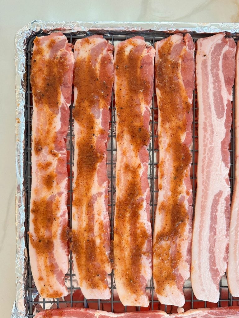 Five uncooked bacon strips on a wire rack, seasoned with spices, on a foil-lined baking sheet.