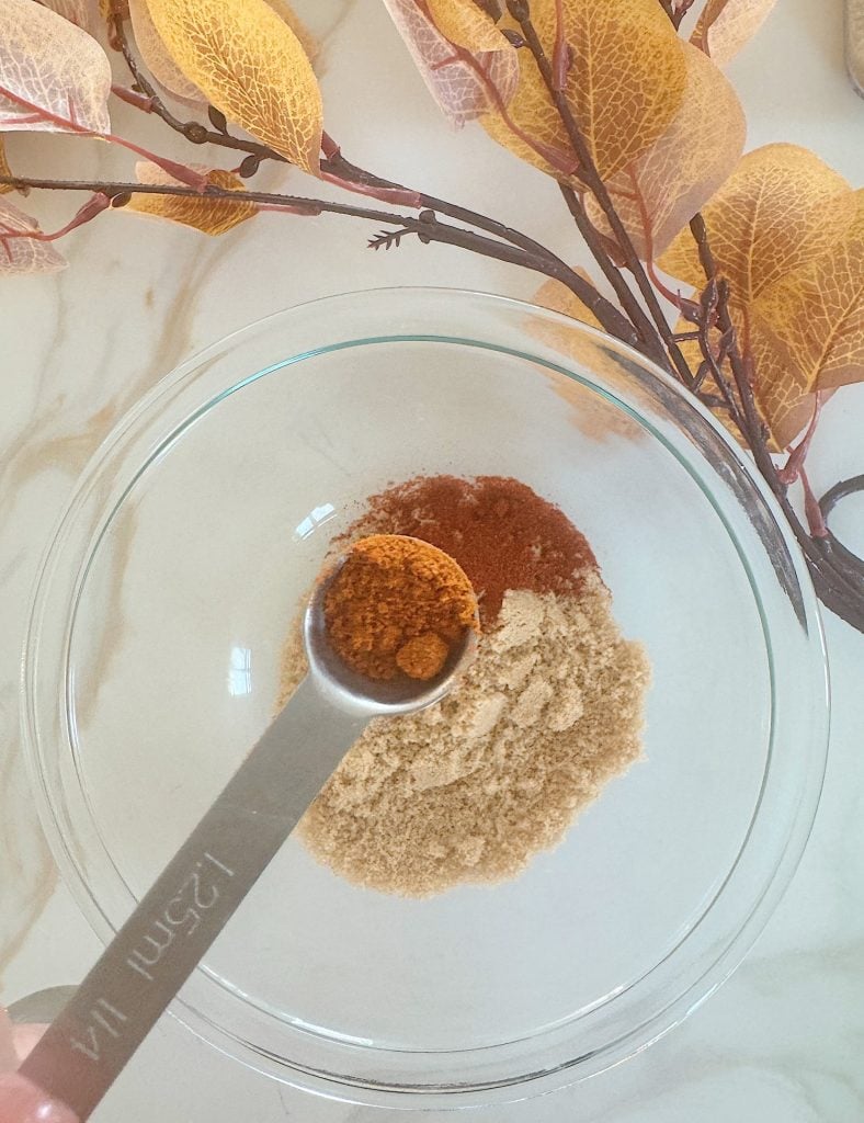 A spoonful of spice being added to a glass bowl containing various spices. Autumn leaves are placed around the bowl on a marble surface.