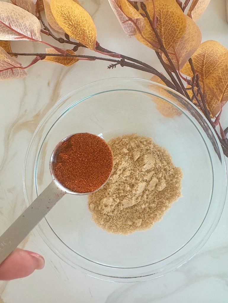 A tablespoon of red spice is held over a bowl containing a beige powder, next to branches with yellow leaves on a marble surface.