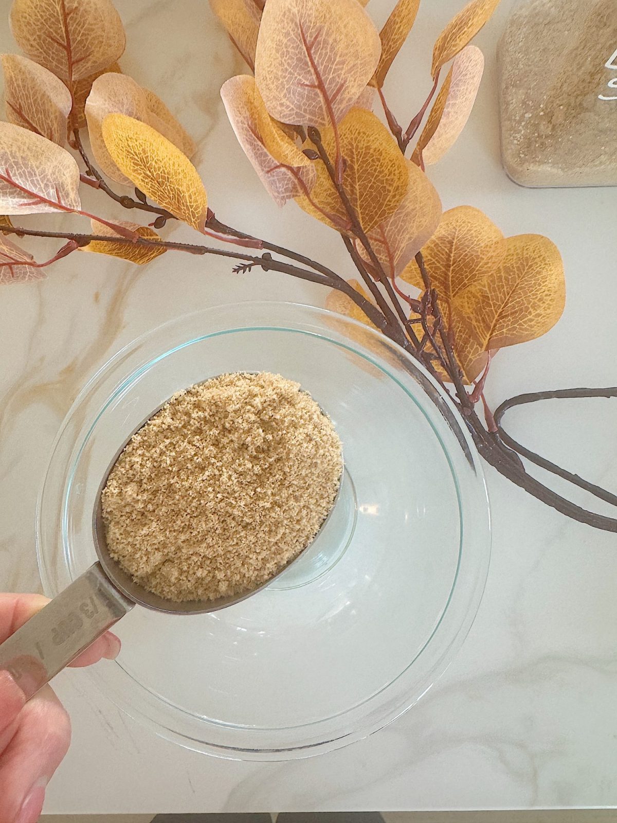 A hand holds a measuring cup of brown sugar over a clear glass bowl. Yellow and brown leaves lie on a light-colored surface in the background.
