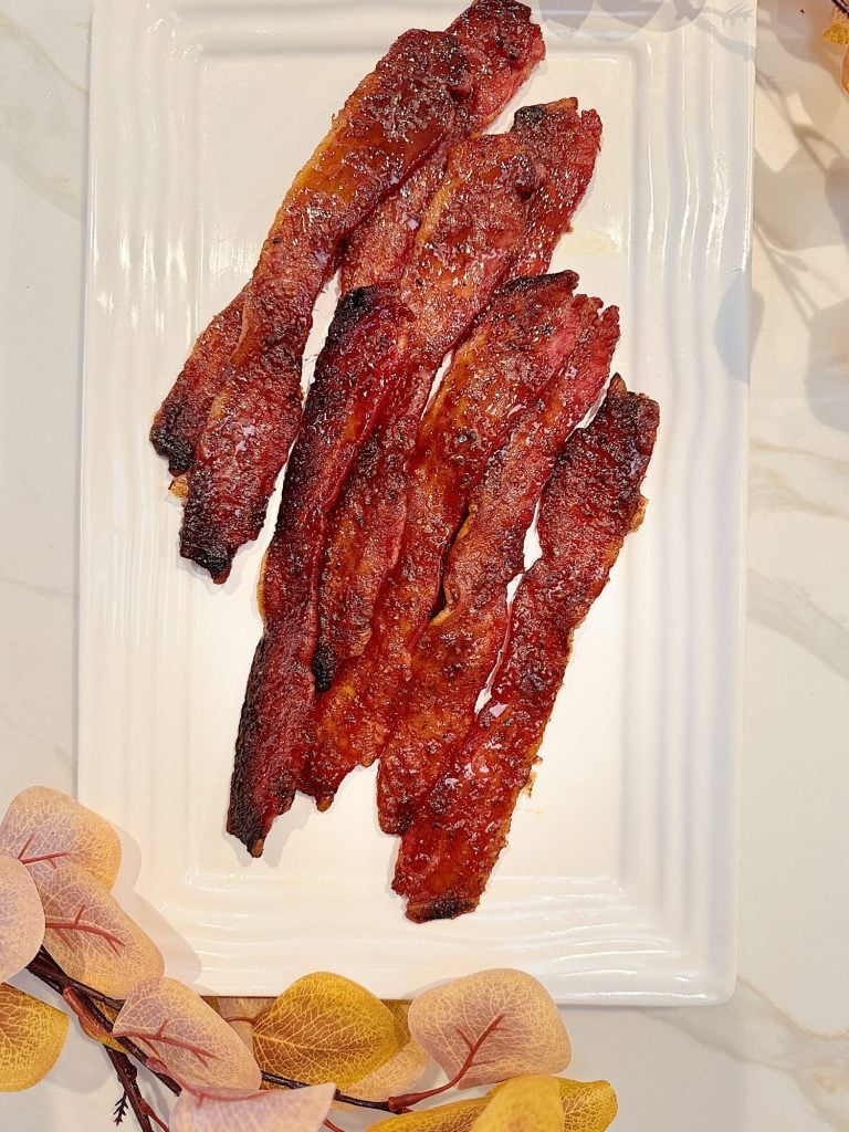 Five strips of cooked bacon on a rectangular white plate, surrounded by decorative leaves.