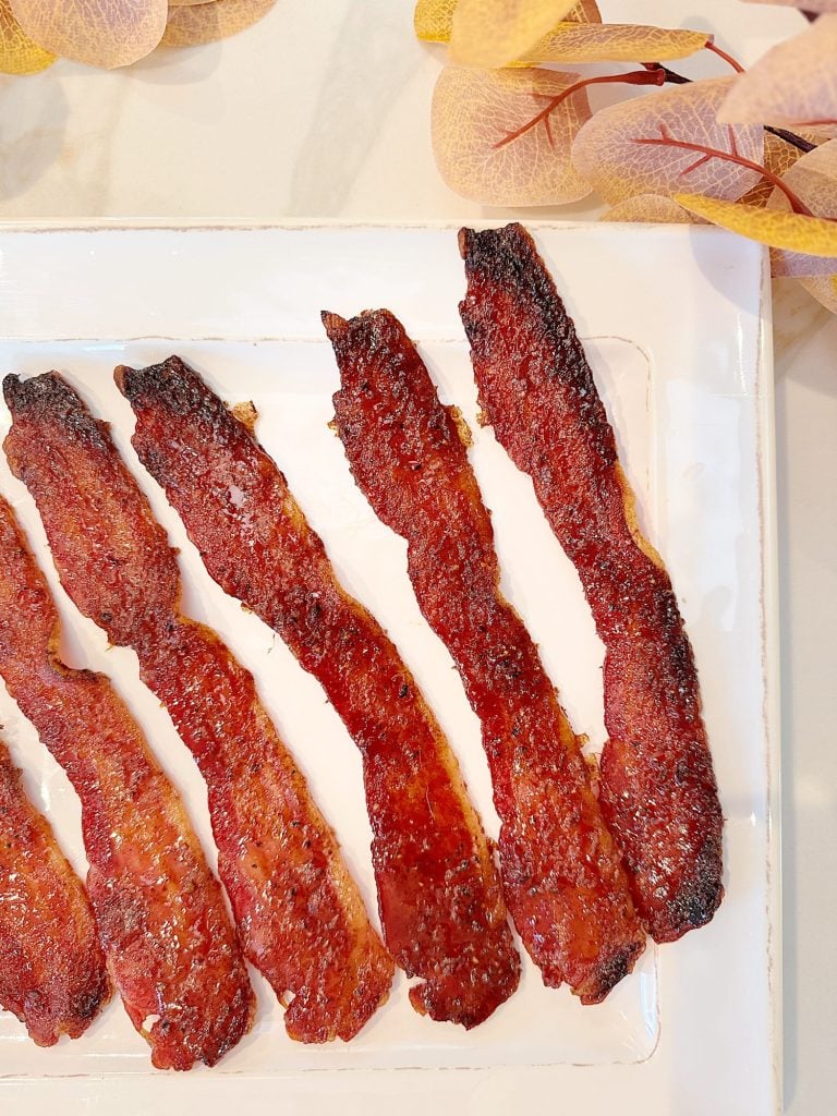 Seven strips of cooked bacon arranged on a white rectangular plate, with decorative leaves in the corner.