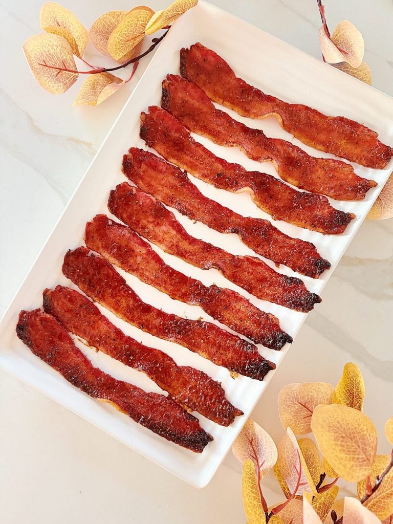 A rectangular white plate with twelve strips of cooked bacon, arranged neatly, with light-colored leaves decorating the corners.