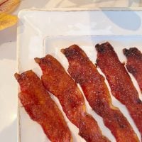 Four strips of crispy bacon on a white rectangular plate, with autumn leaves in the background.