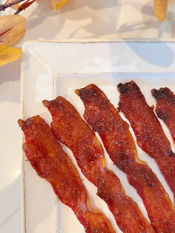 Four strips of crispy bacon on a white rectangular plate, with autumn leaves in the background.