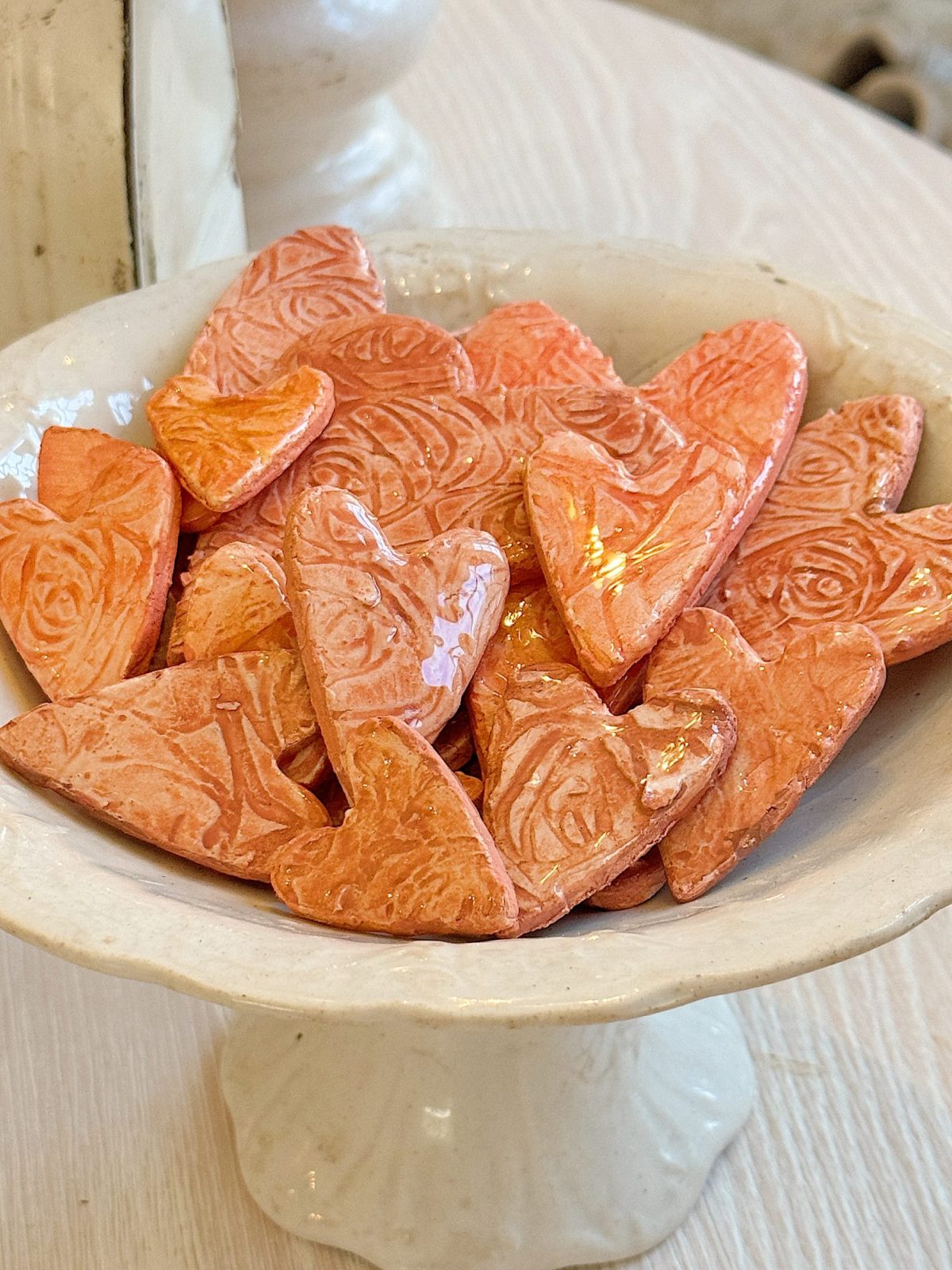 A white pedestal bowl filled with heart-shaped pink ceramic pieces with an embossed floral pattern.