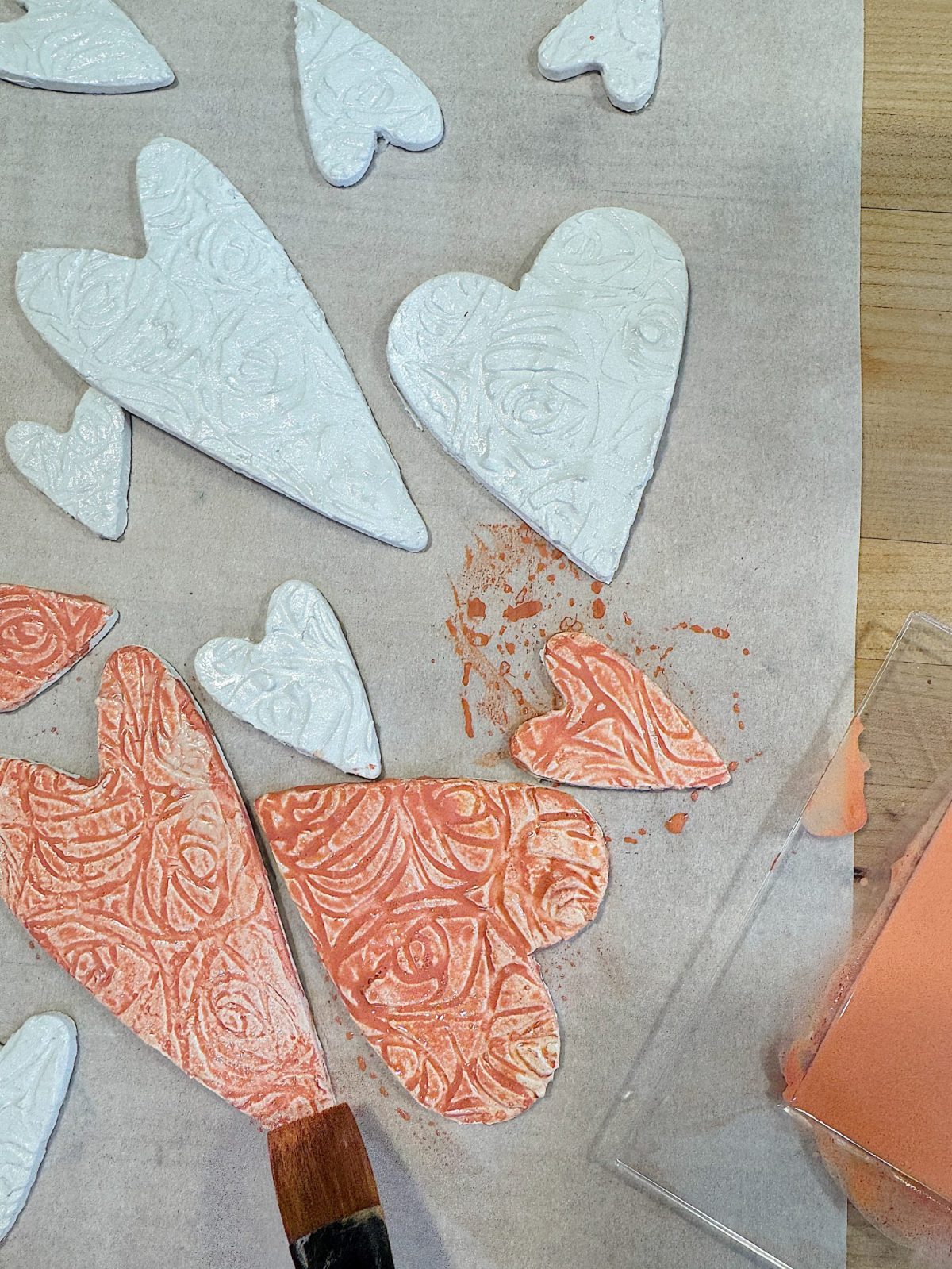 Heart-shaped clay pieces with embossed patterns being painted. Some are white, others are coral. A paintbrush is visible, along with a splatter and a tray of paint.