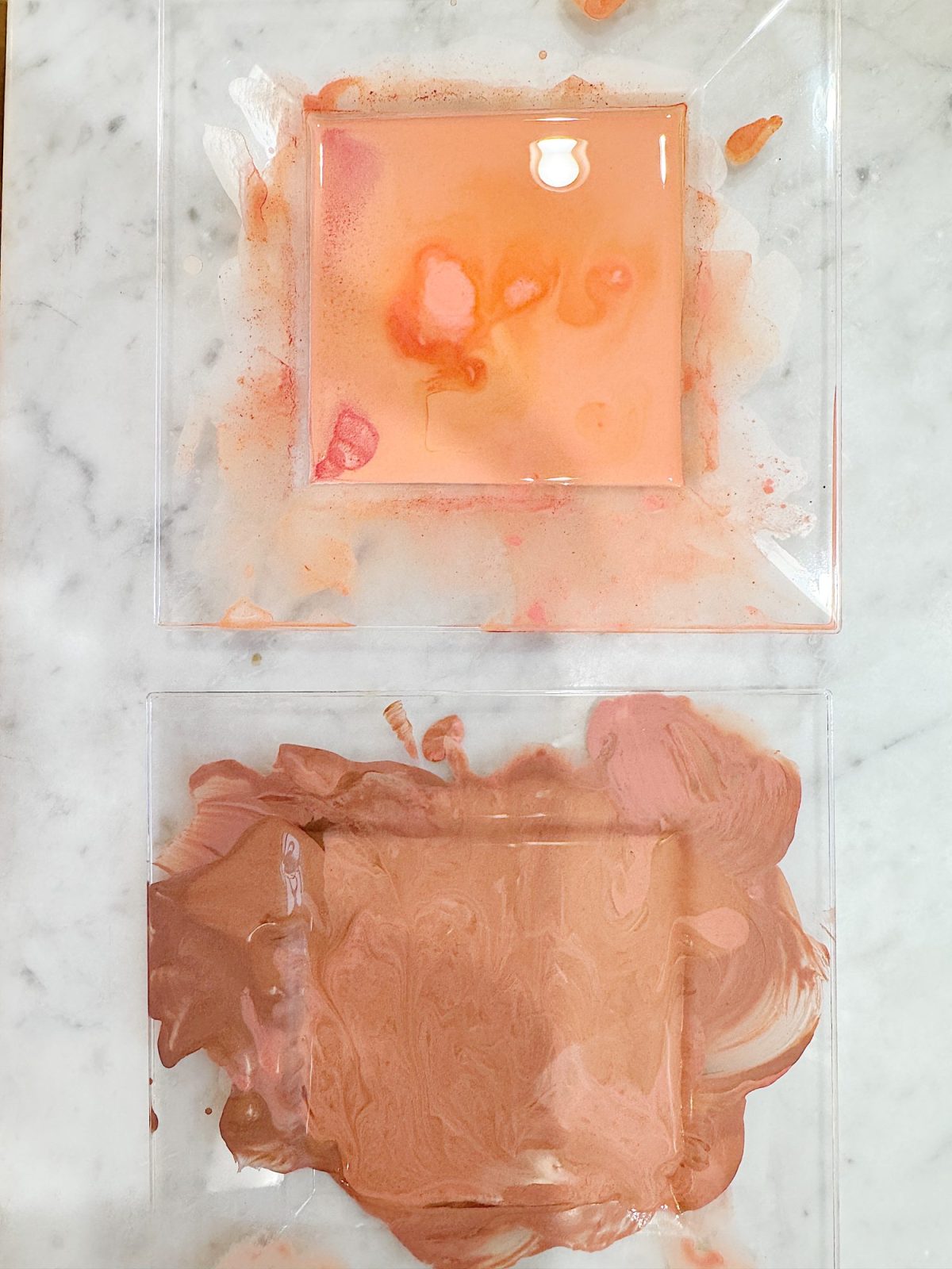 Two transparent square dishes containing swirled paint in shades of pink, peach, and brown on a marble surface.