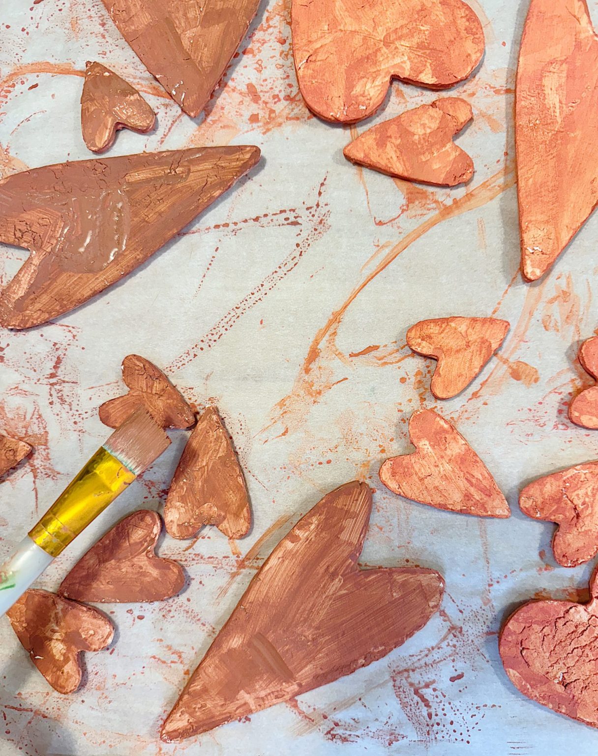 Painted clay hearts of varying sizes in reddish tones scattered on paper; a paintbrush applies finishing touches.