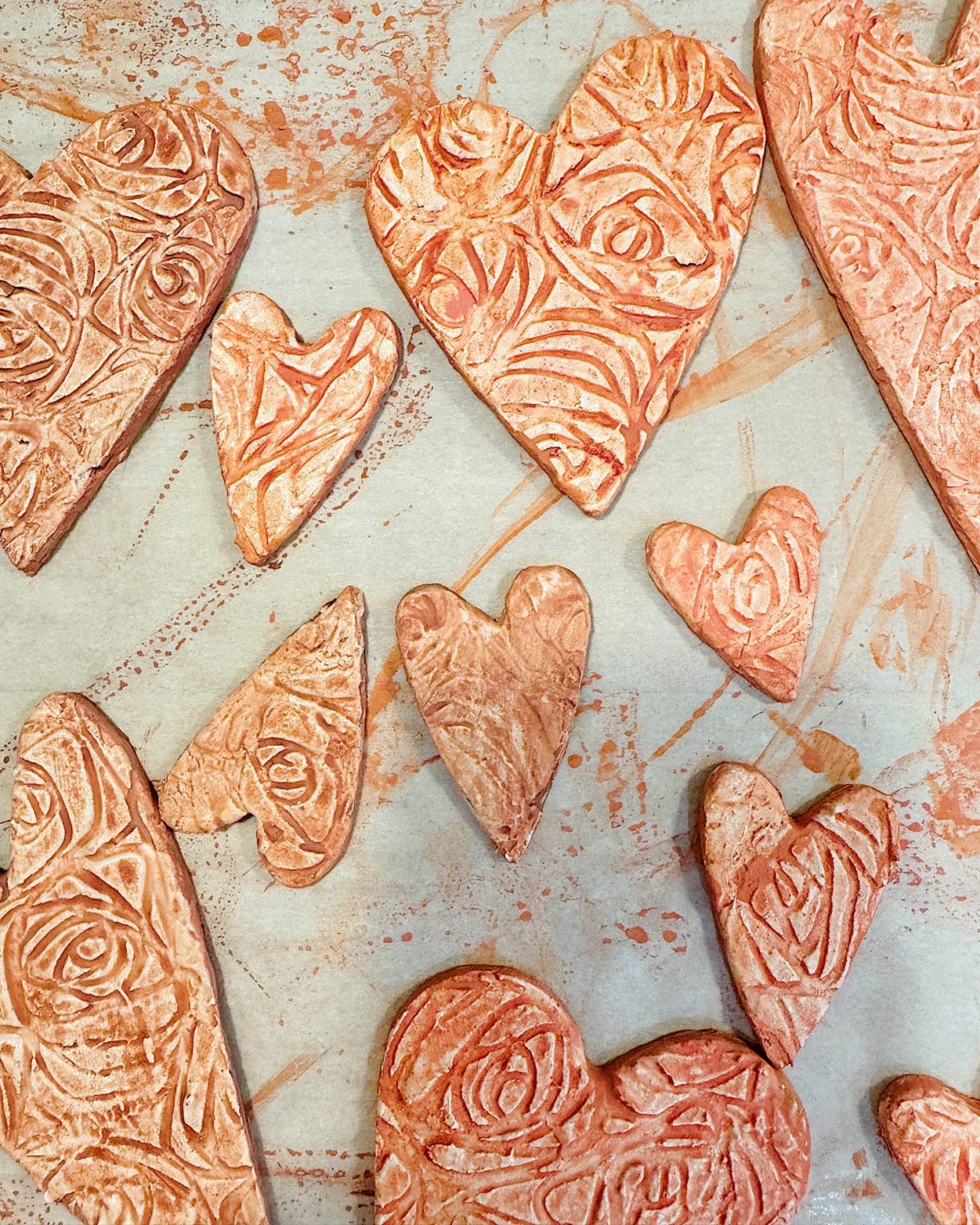 Heart-shaped cookies with rose-patterned designs are arranged on a parchment-lined surface, featuring variations in size and a textured appearance.