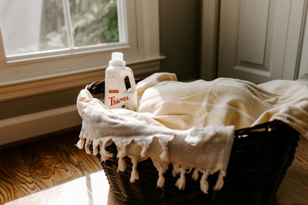 A bottle of Thieves laundry detergent rests in a wicker basket lined with a beige towel, positioned on a wooden floor near a sunlit window.