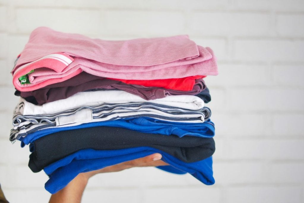 A hand holding a stack of neatly folded clothes in various colors, including pink, red, white, and blue, against a white brick wall background.