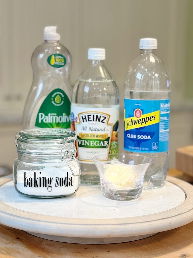 A display of household cleaning items including a jar of baking soda, Palmolive dish soap, Heinz vinegar, Schweppes club soda, and a small dish of butter on a round tray.