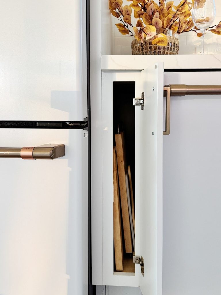 A small kitchen cabinet is open, revealing cutting boards stored inside. A refrigerator and decorative vase with orange leaves are visible nearby.