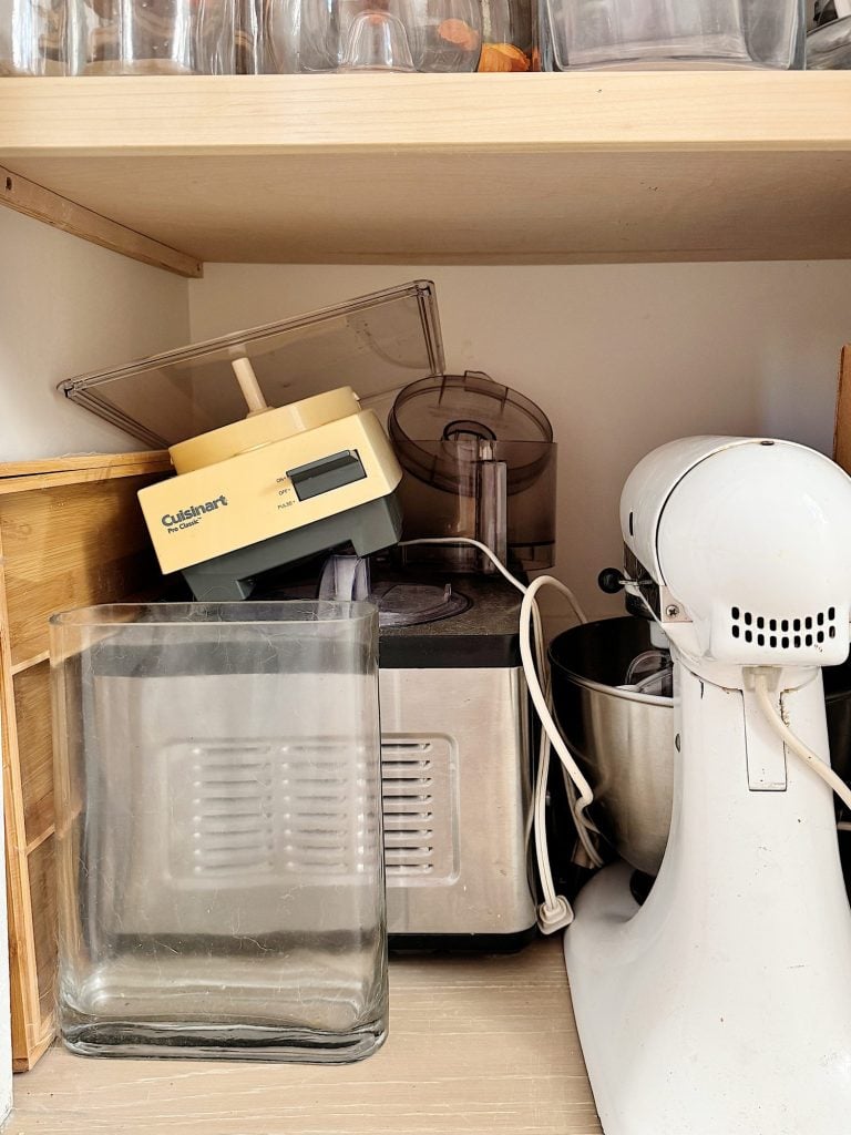 A shelf with kitchen appliances including a white stand mixer, a food processor, and a blender with a yellow base. Empty glass jars are also present.