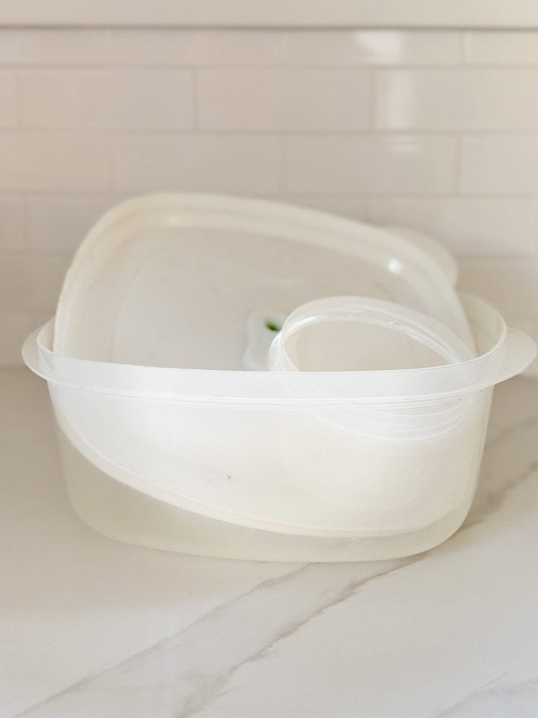 Stacked transparent plastic food containers with lids on a marble counter, against a white tiled backsplash.