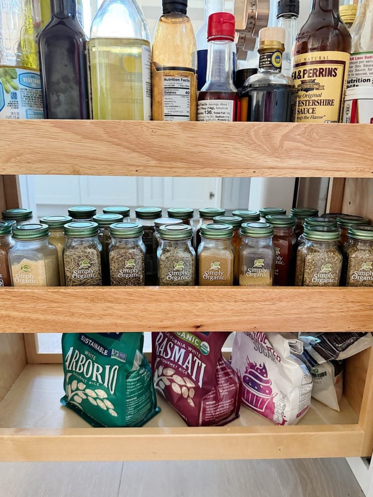 A pantry shelf with bottles of oils and sauces on top, a row of spices in the middle, and bags of rice and flour on the bottom.