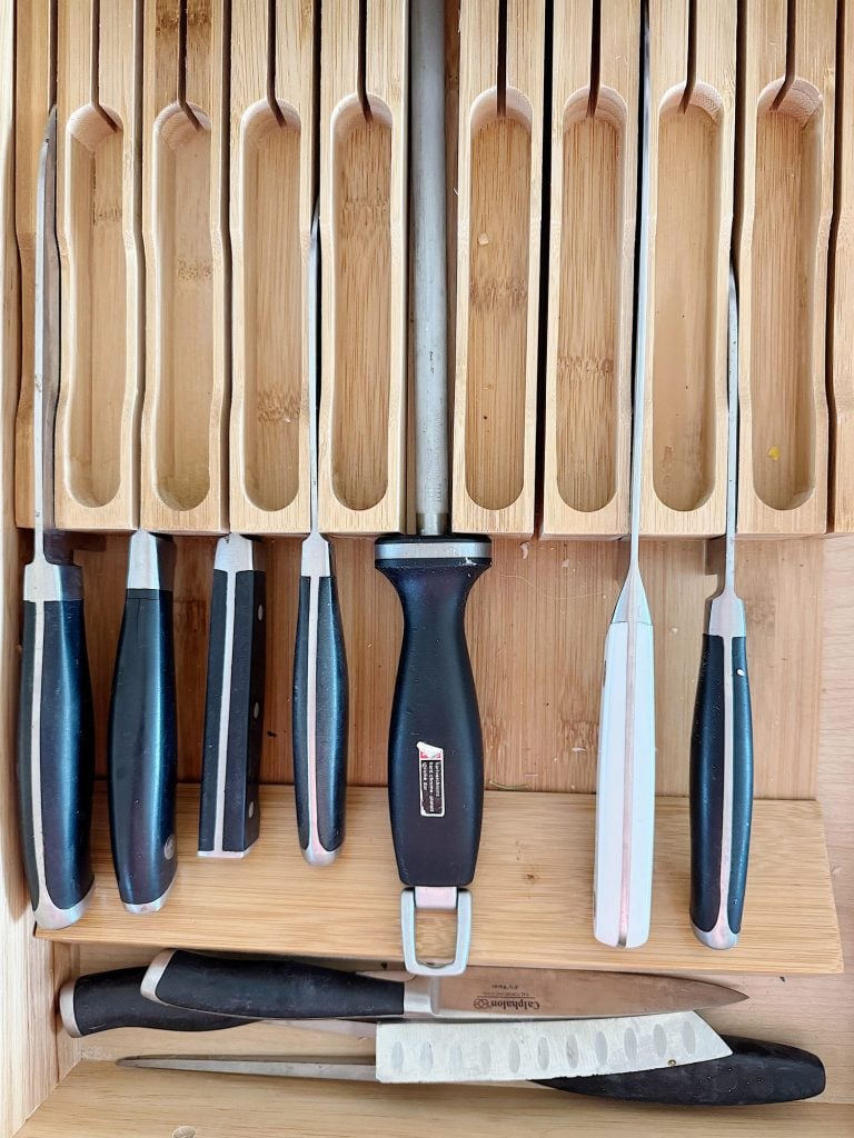 A drawer organizer holding seven various knives and a honing rod with black and silver handles. A separate smaller knife sits at the bottom.