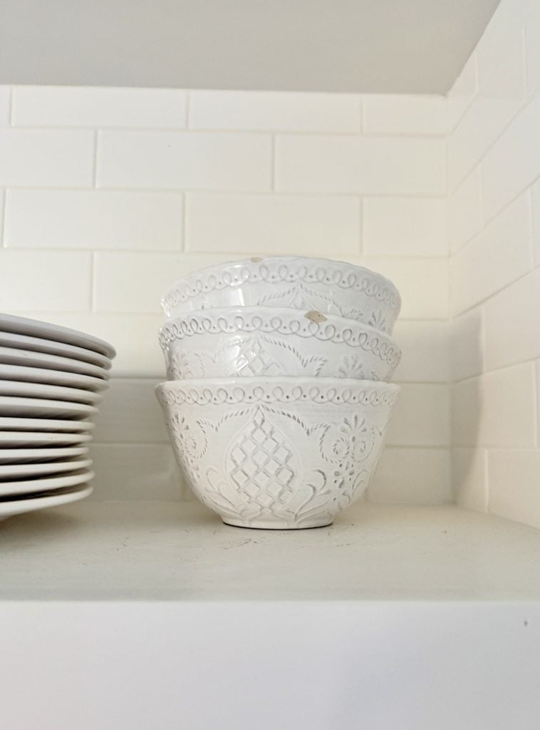 Stacked white ceramic bowls with patterns, placed beside a stack of white plates on a kitchen shelf with a white tiled backsplash.