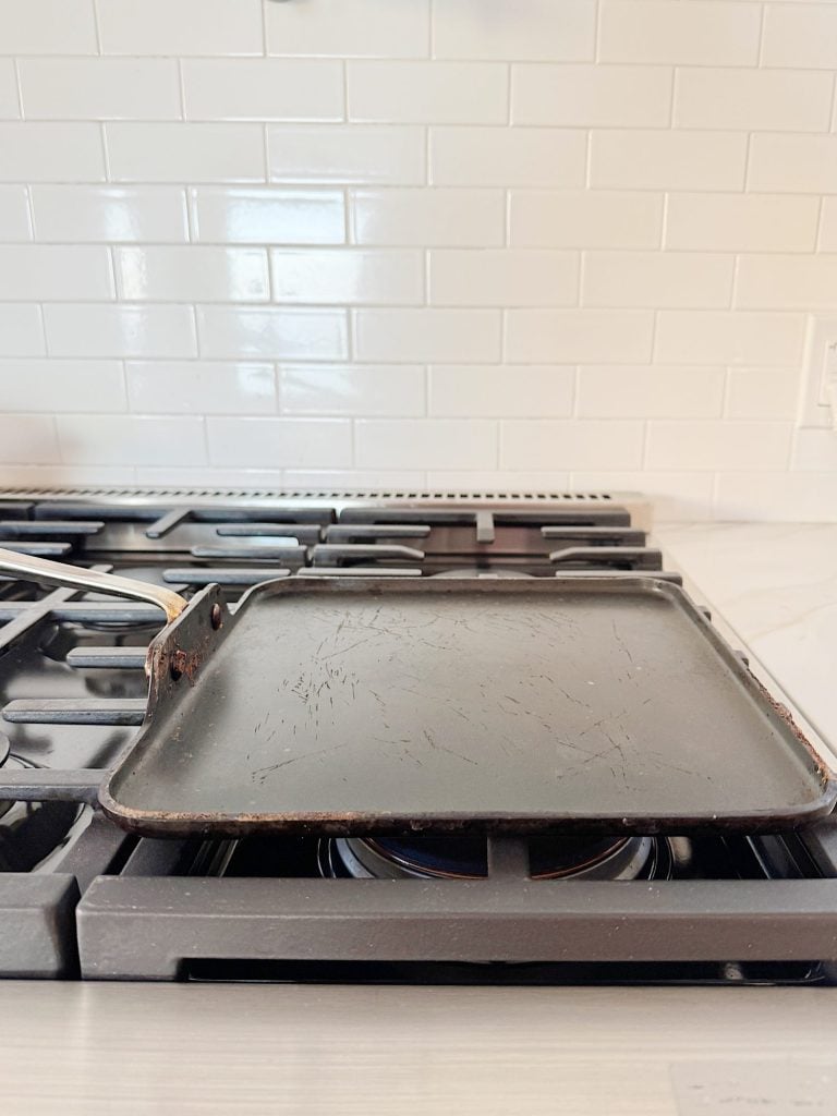 A well-used griddle on a stove with a white tile backsplash.