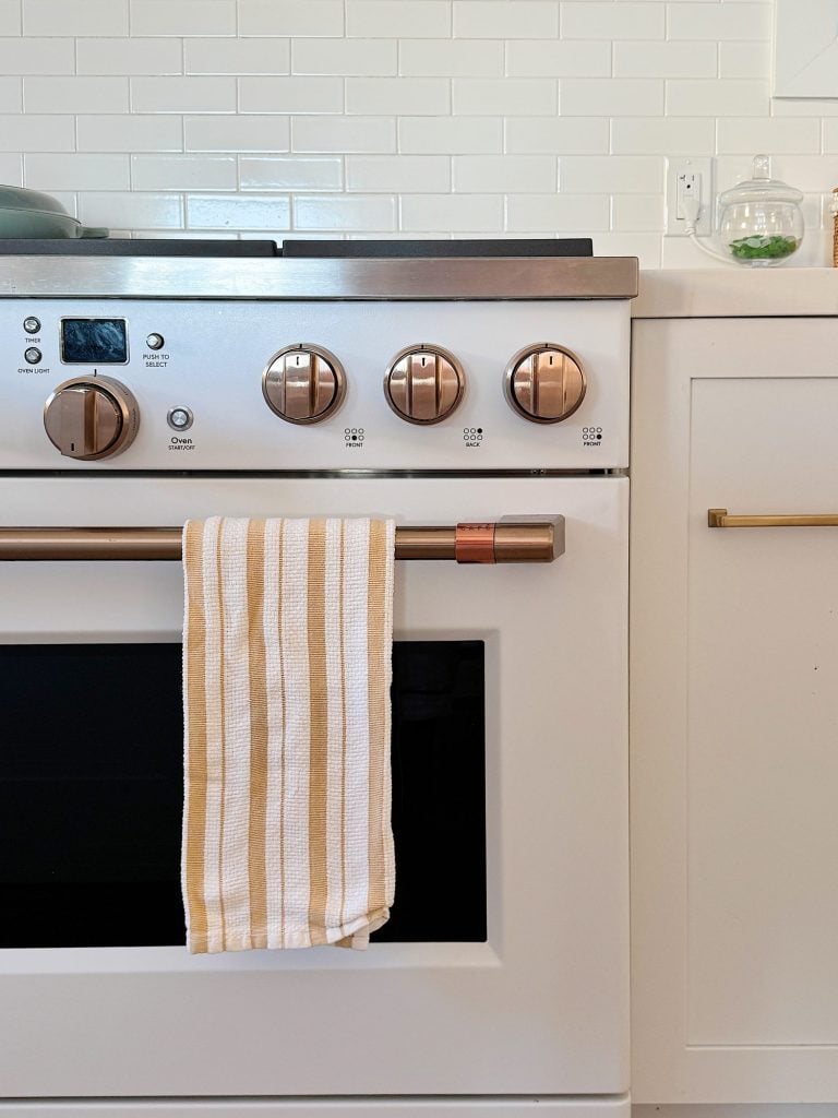 A white stove with copper knobs and a matching oven handle. A beige and white striped towel hangs from the handle. White tiles and cabinetry are in the background.