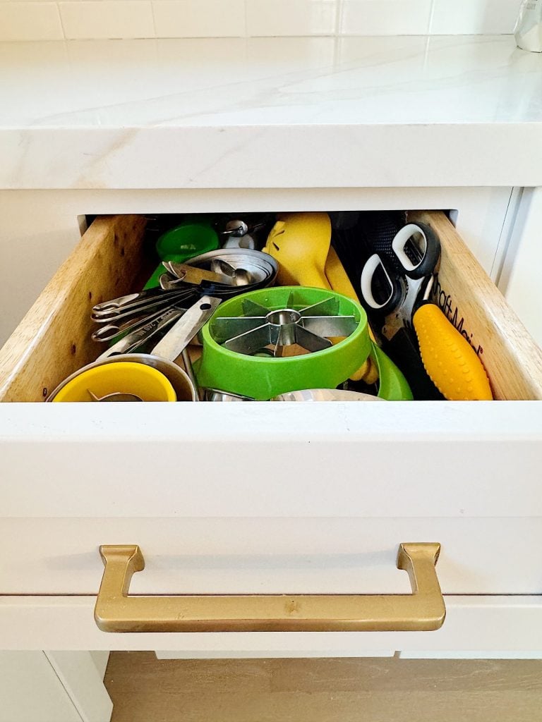 Open kitchen drawer filled with assorted utensils, including a green apple slicer, metal measuring spoons, scissors, and a yellow brush.