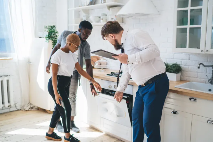 Three people in a kitchen, two examining an oven while one holds a clipboard, in a modern white interior.