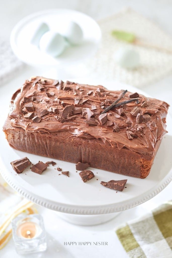 A chocolate loaf cake with chocolate frosting and chocolate chunks on top, placed on a white cake stand.