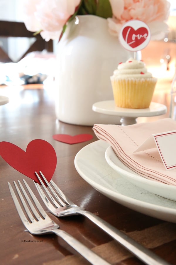 Table setting with heart-shaped decor, a cupcake on a stand, pink napkin, and cutlery on a white plate.